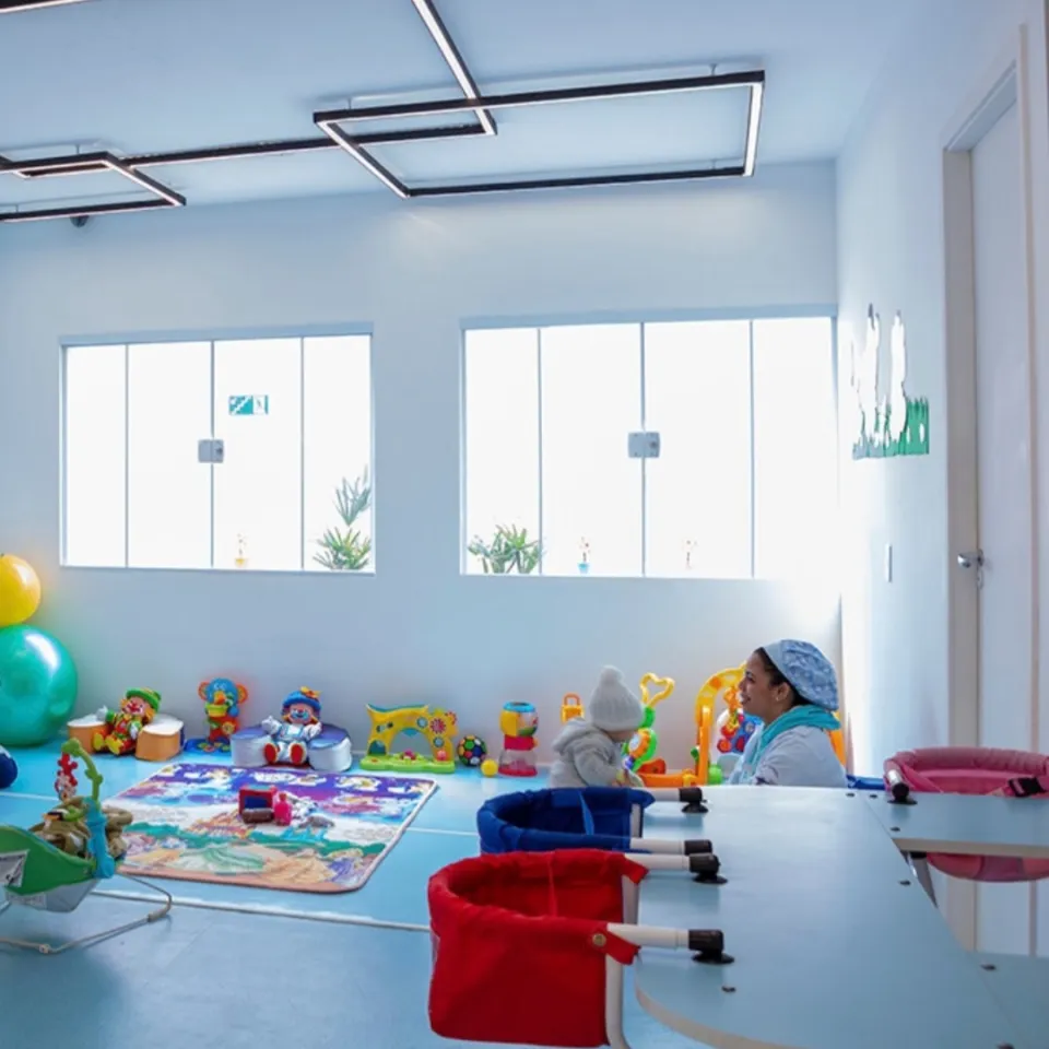 Sala de berçário da Baby Nurse, com paredes azul-claro, iluminação natural, brinquedos educativos, e espaços preparados para bebês. Uma educadora acompanha um bebê durante uma atividade.