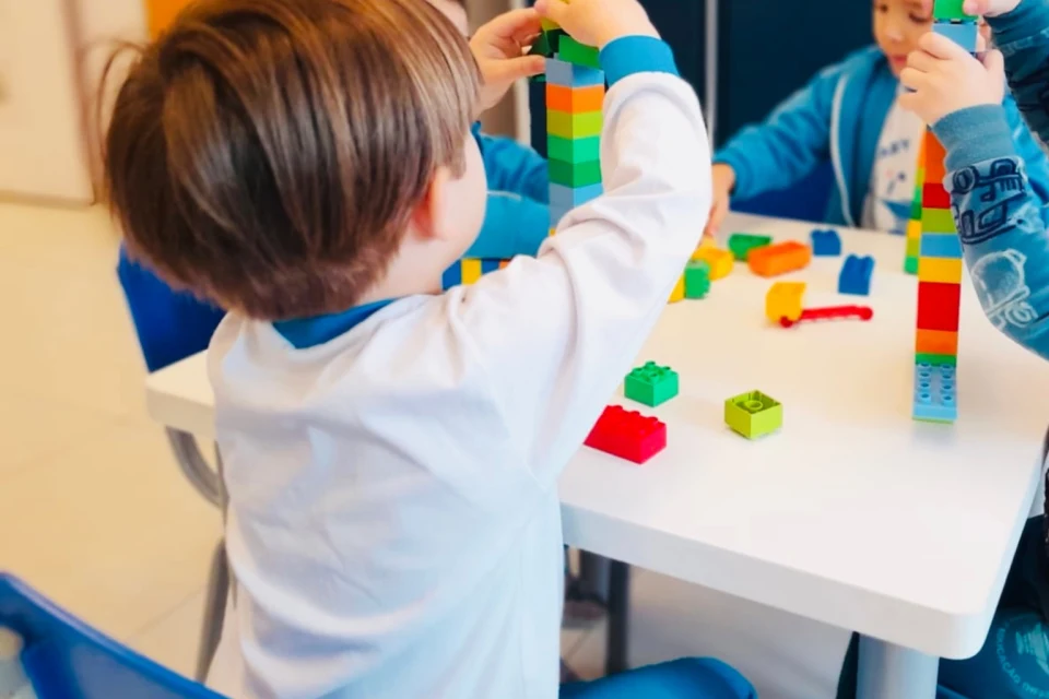 Crianças pequenas brincam juntas em uma sala de Educação Infantil, construindo torres com blocos coloridos sobre uma mesa branca. A atividade estimula a criatividade, a coordenação motora e a interação social.